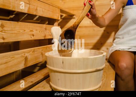 hand of unknown caucasian woman in sauna spa taking water from bucket with wooden spoon to pour on the hot stones Stock Photo