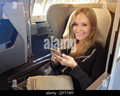 First class is the only way to go. Portrait of a young woman using her cellphone while sitting in first class in an airplane. Stock Photo