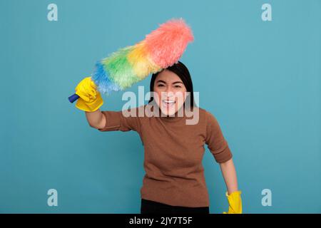 Angry energetic young asian houseworker holding colorful dust brush in yellow glove isolated against a blue background, Cleaning home concept, Crying female with face expression Stock Photo