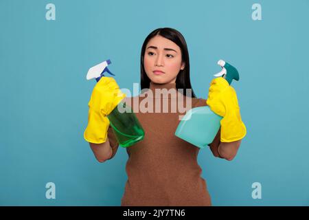 Asian tired housewife frustrated from overwork holding two detergent sprayers in yellow gloves isolated on a blue background, Housekeeping and hygiene concept, Daily homework Stock Photo