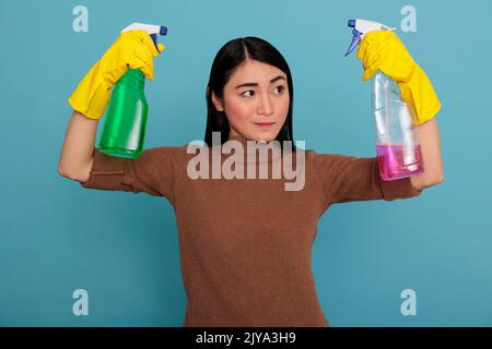 Strong serious asian housemaid raising arms from chores holding two detergent sprays in the yellow gloves isolated on a blue background, Cleaning home concept, Energetic confidence woman at work Stock Photo