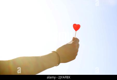 An outstretched hand in bright sunlight holding a deep red love heart shaped lollypop, lollipop, candy stick towards the sky. Stock Photo