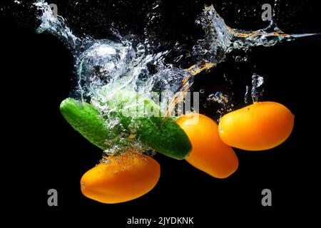 Fresh green cucumbers and yellow plum tomatoes dropped in water with splashes against black background. Stock Photo