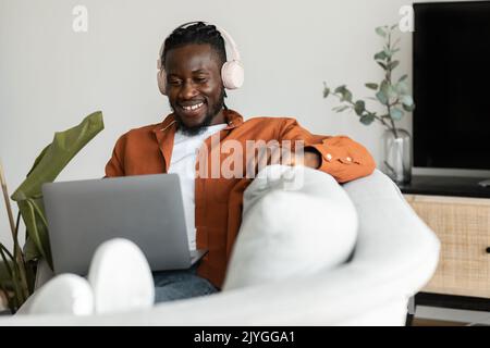 Positive black freelancer man in headphones working on laptop at home, sitting on couch in living room, free space Stock Photo