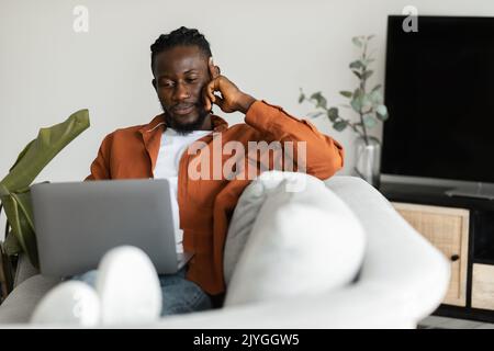 Focused african american freelancer man working on laptop computer at home, serious black male sitting on sofa Stock Photo