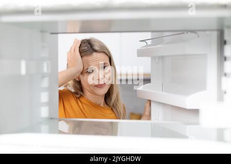 Sad blonde woman looking inside open empty fridge Stock Photo