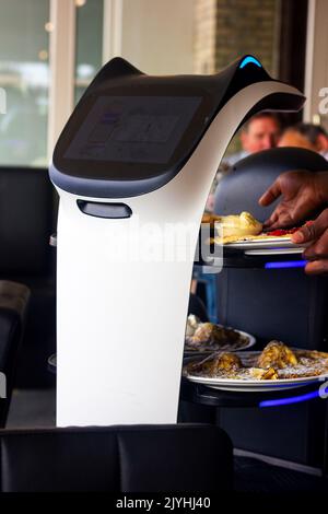 A close portrait of a serving robot on wheels with a screen displaying a cute cat face, riding through a restaurant with plates with pancakes on its t Stock Photo