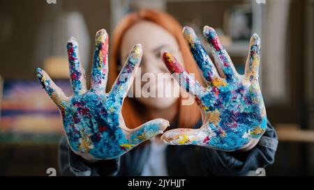 Happy funny Caucasian red-haired woman in eyeglasses girl painter wear glasses looking at camera posing in art studio laughing show palms dirty soiled Stock Photo