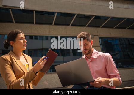 A female business worker is reading the notes to her male colleague and he is writing them down on the lap top Stock Photo