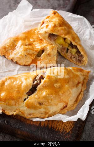 Traditional Cornish pasty filled with beef meat, potato and vegetables closeup on the paper on the wooden board. Vertical Stock Photo