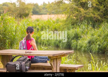 Rear view of young woman with ponytail wearing sports cap, relaxing, sitting on picnic bench overlooking countryside canal on a warm, summer day. Stock Photo
