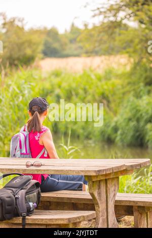 Rear view of young woman with ponytail wearing sports cap, sitting on a picnic bench in the countryside looking at a UK canal during warm  summertime. Stock Photo