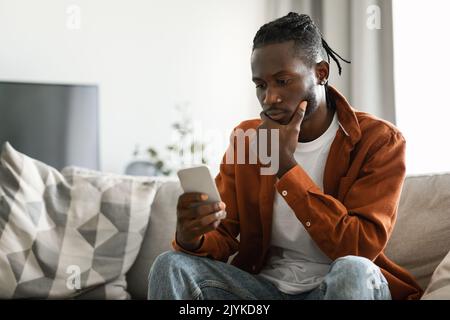 Pensive african american man using smartphone, sitting on sofa and looking at phone with serious expression, free space Stock Photo