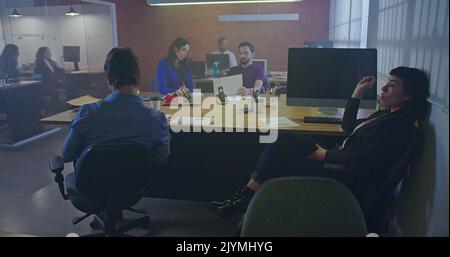 People working at Office workplace interior at night. Interior of busy employees in a coworking space. Corporate businessmen and businesswomen sitting Stock Photo