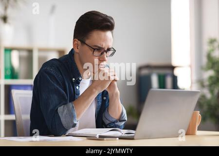 Thougthful Young Businessman Sitting At Workplace And Looking At Laptop Screen Stock Photo