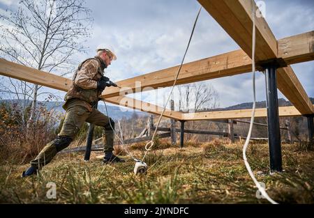 Man building wooden frame house on pile foundation. Side view of male worker drilling hole by electric drill in timber framing of future house on construction site. Builder wearing white helmet. Stock Photo
