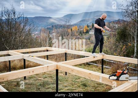 Man worker building wooden frame house on pile foundation in the mountains. Carpenter measuring wooden planks. Carpentry concept. Stock Photo