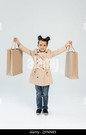 full length of cheerful kid in trench coat and jeans holding shopping bags on grey, black friday concept,stock image Stock Photo