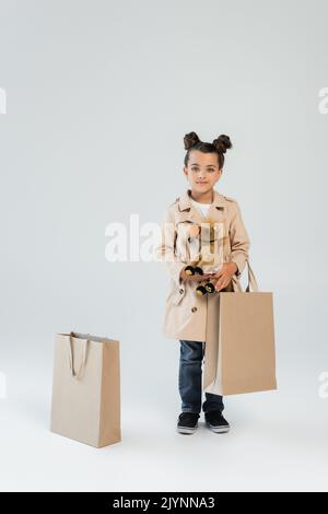 full length of cheerful kid in trench coat and jeans holding soft toy and shopping bag on grey,stock image Stock Photo