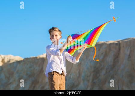 A handsome boy in a white shirt with a multicolored kite on blue sky background. Stock Photo