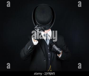 Portrait of English Gentleman in Dark Suit and Leather Gloves Tipping Bowler Hat in Greeting. Classic British Butler or Businessman. Stock Photo