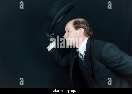 Portrait of British Businessman in Black Suit and Leather Gloves Tipping Bowler Hat and Bowing in Polite Greeting. Classic English Gentleman Stock Photo
