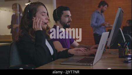 Telemarketers communicating with clients wearing headsets in front of laptops. Female and male employees working inside office workplace speaking with Stock Photo