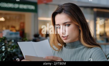 Portrait of beautiful young caucasian millennial woman recipient girl sitting in cafe receiving letter opens envelope reads test results bank notice Stock Photo
