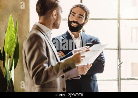 Two businessman colleagues discussing a project standing in the office against a big window, business lifestyle concept Stock Photo