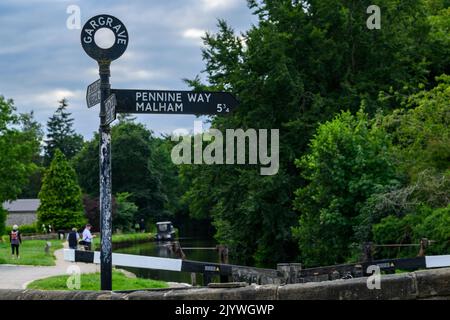 Waymark sign pointing to Pennine Way footpath route by canal lock & people walking - Leeds and Liverpool Canal, Gargrave, North Yorkshire, England UK. Stock Photo