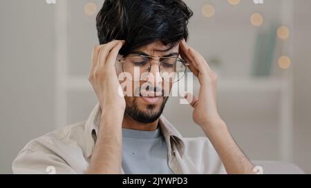 Close-up arabic man bearded guy with glasses feels stress overwork cranial pressure massages weight on head suffering from headache migraine sighing Stock Photo