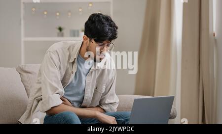 Young hispanic arab indian business man freelancer worker male bearded student wearing glasses sitting at home office on sofa with laptop feels Stock Photo