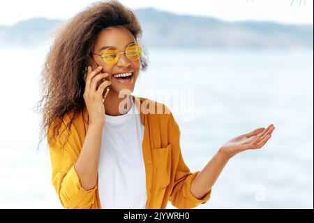 Joyful curly beautiful African American stylish girl, in an orange shirt and with orange glasses, stands outdoors, talking on a mobile phone, gesturing with her hand, looking away, smiling Stock Photo