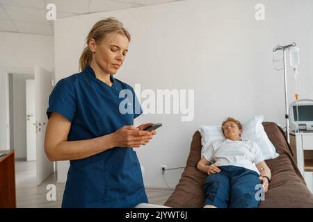 Nurse in uniform using phone standing near sick patient in hospital ward Stock Photo