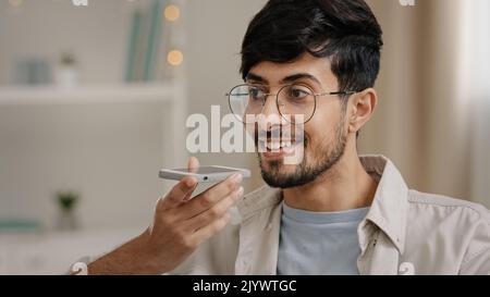 Friendly young arab man recording voice recognition message on speakerphone sitting at home. Bearded guy with glasses using virtual assistant app sets Stock Photo
