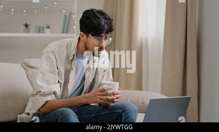 Young freelancer male business man bearded guy in glasses drinks tea coffee delicious drinking at home sitting on couch watching movie in laptop Stock Photo