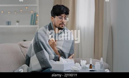 Young sick guy arabic man with glasses sits at home sofa covered with blanket plaid suffers from cold illness seasonal allergy virus blowing runny Stock Photo