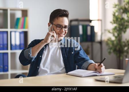 Millennial man writing notes while making phone call at workplace in office Stock Photo