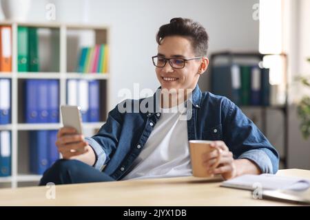 Young Male Office Employee Relaxing With Smartphone And Coffee At Workplace, Stock Photo