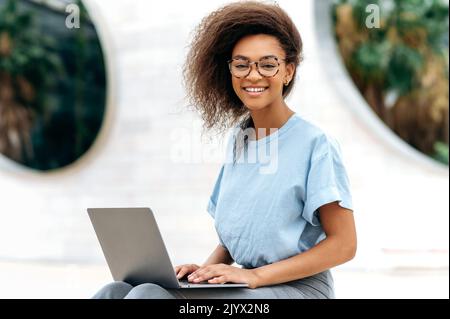 Photo of an elegant lovely positive confident African American woman, with glasses, manager, company seo, freelancer, sits with laptop outdoors near business center, looks at camera, smiles friendly Stock Photo