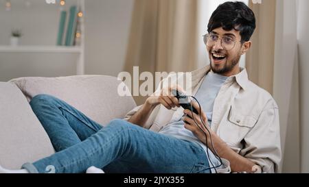 Arab guy bearded man with glasses lying on couch at home playing console online video game competition emotionally winning yes hand gesture victory Stock Photo