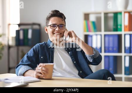 Handsome Young Man Enjoying Phone Conversation During Coffee Break In Office Stock Photo