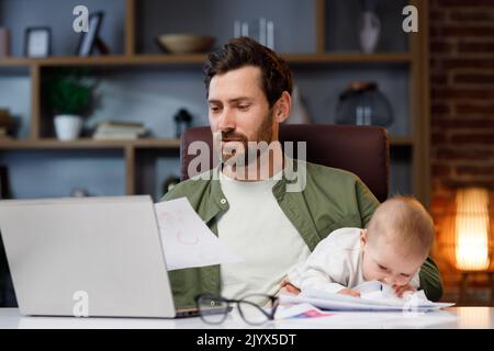 Father on maternity leave, holding an online conference, a meeting in the home office with a small child in his arms. Dad with a child. Freelancer Stock Photo