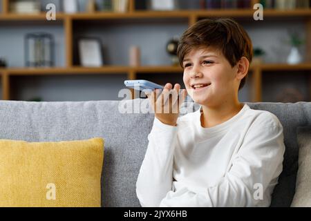 Young cute boy recording voice message on smartphone. Smiling teenager using virtual assistant app sitting at coach at home. Voice recognition, audio Stock Photo