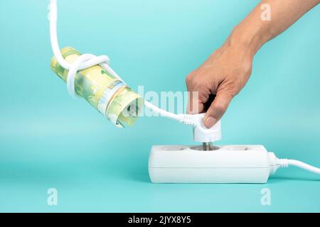 Woman hand holding electric power plug with Euro banknote on cord and plugging it in power strip extension cord on blue background. Electricity cost Stock Photo