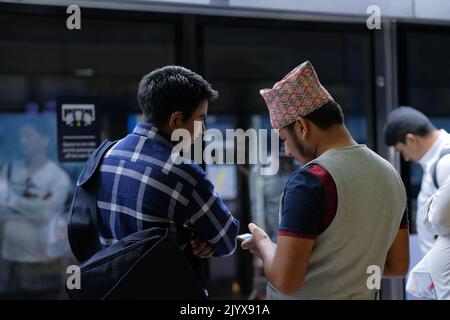 Two men talk while waiting at a train platform. One man is wearing a vest and an embroidered Islamic hat. Other male has a large black bag slung over. Stock Photo
