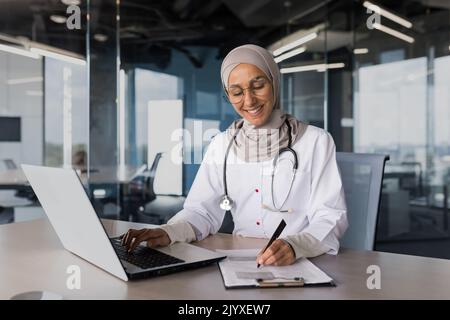 Arab muslim woman in hijab doctor working in modern clinic office with laptop, doctor on paper work in white medical coat with stethoscope and glasses. Stock Photo