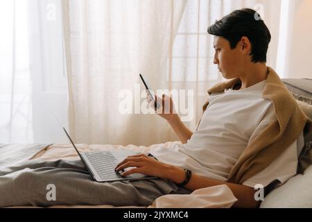 Asian boy using cellphone and working with laptop while resting on bed at home Stock Photo