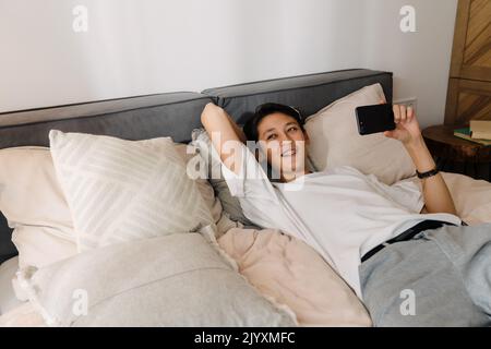 Asian boy smiling and using mobile phone while resting on bed at home Stock Photo