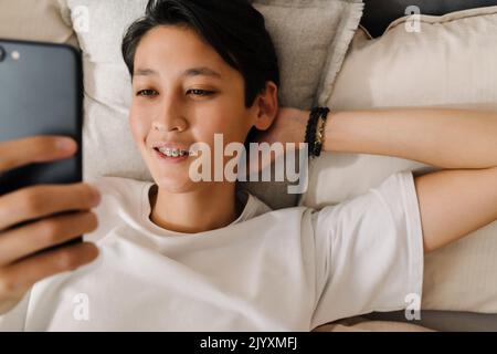 Asian boy smiling and using mobile phone while resting on bed at home Stock Photo